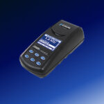 A black handheld electronic device with a digital screen and blue buttons is shown against a blue gradient background. The screen displays measurement data and the brand name Hach.