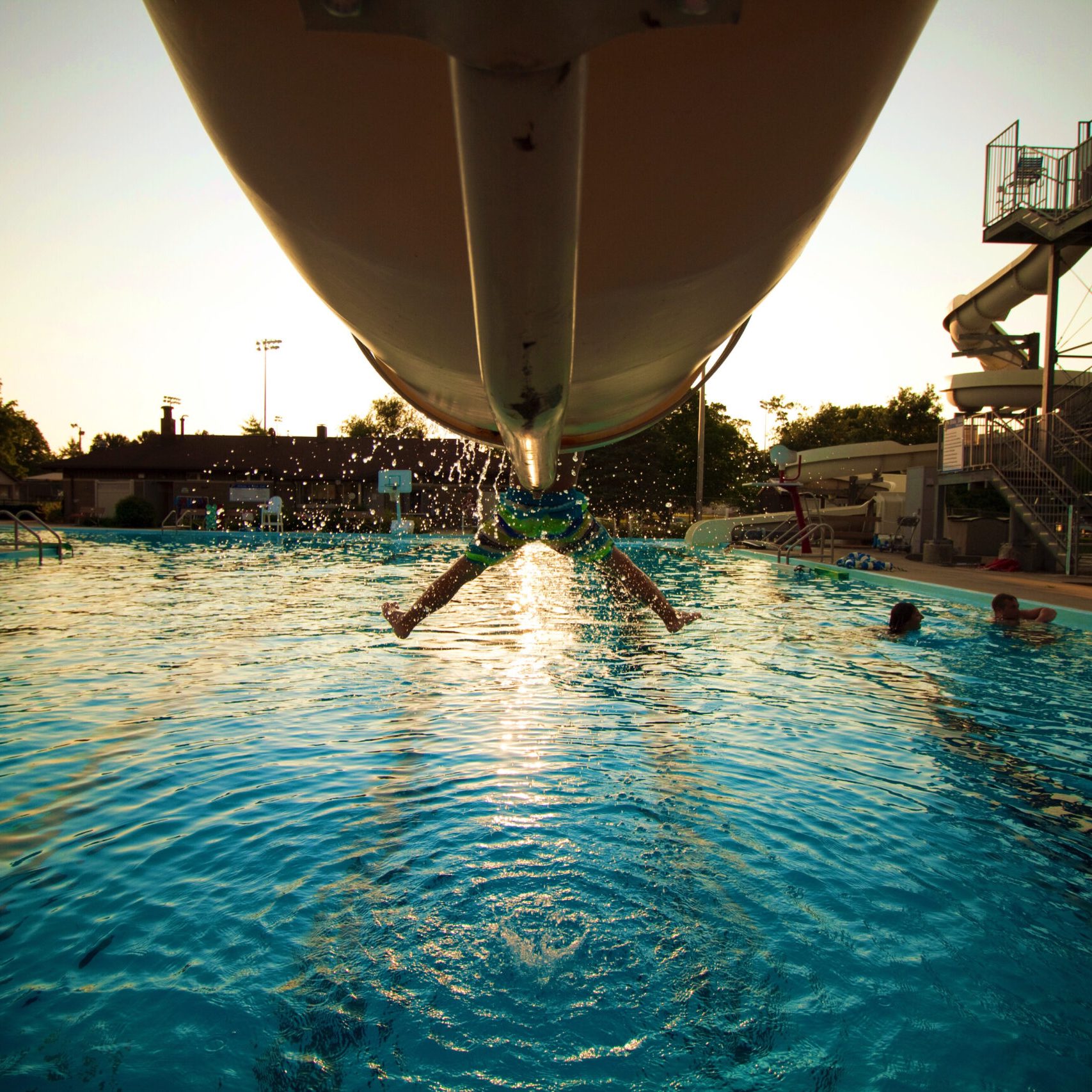 A person comes down a waterslide and splashes into a swimming pool, with water droplets in midair and other swimmers nearby. The sun is setting, casting a warm glow over the scene.